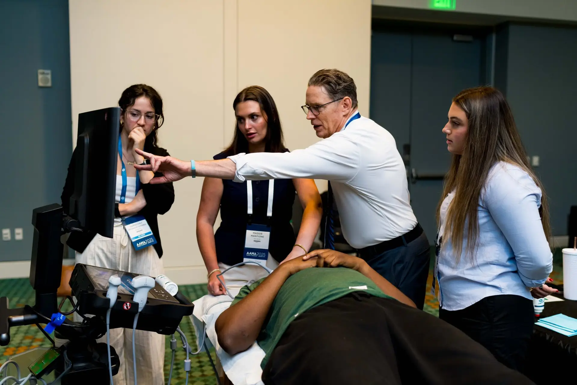Instructor leads a training for three women beside a patient on an exam table in a conference room.