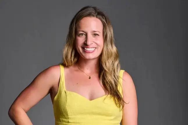 Smiling woman with long blonde hair in a yellow dress, pictured against a plain gray background.