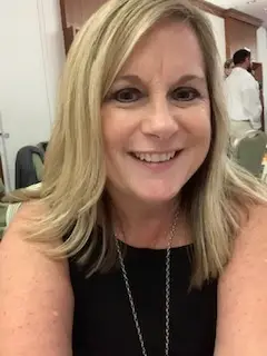 Smiling woman with blonde hair in a black top seated at an indoor professional event.