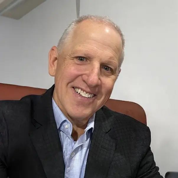 Smiling older man with short gray hair in a dark blazer and blue shirt sits in a chair, facing the camera.