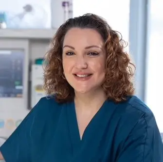 Smiling woman with curly brown hair in blue scrubs sits in a hospital setting with medical equipment behind her.