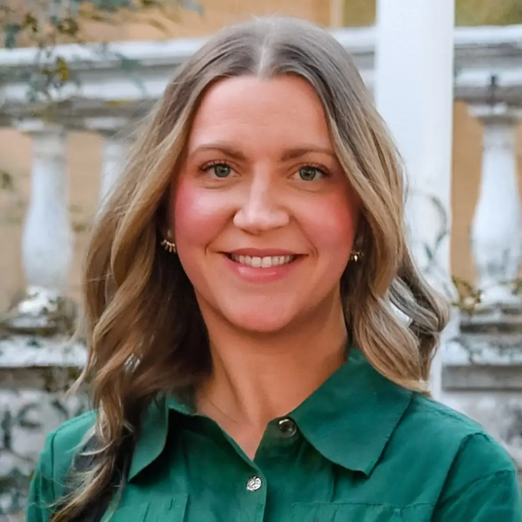 Smiling woman with wavy light brown hair in a green shirt stands outside by a white stone railing.
