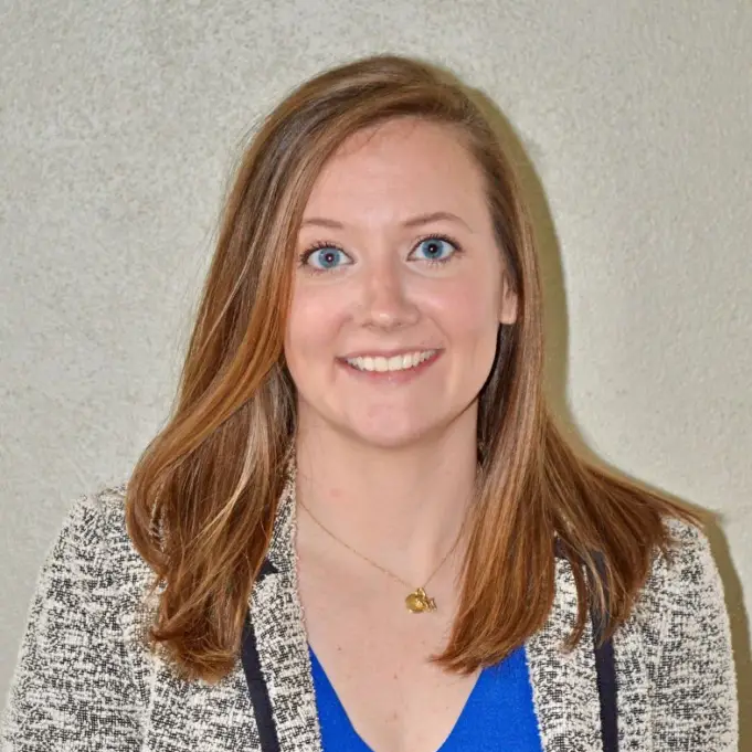 Smiling woman with straight light brown hair in a gray blazer and blue top, wearing a gold necklace, against a light background.