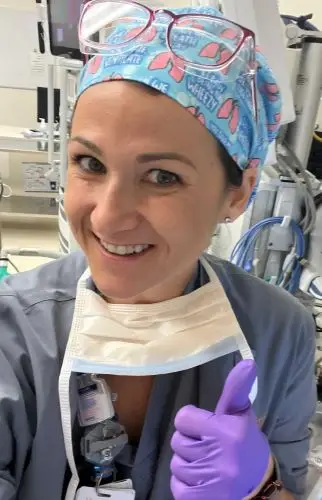 Smiling healthcare worker in scrubs, colorful cap, and purple gloves gives a thumbs up in a medical setting.