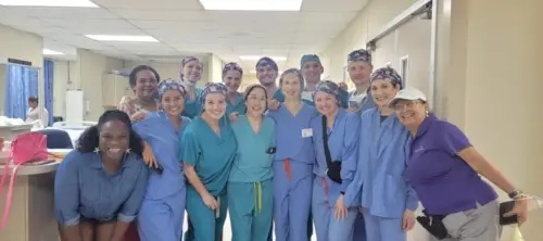 Smiling healthcare workers in scrubs and caps pose in a hospital hallway, joined by two casually dressed colleagues at each end.