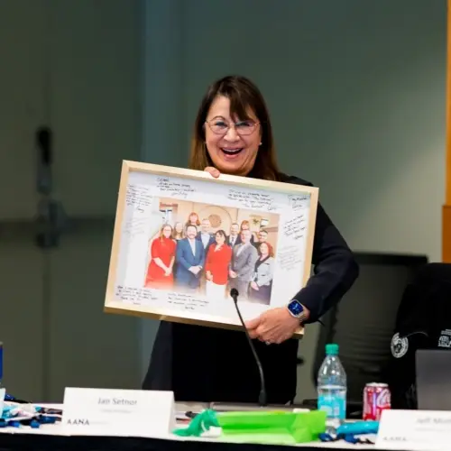 Jan Setnor smiles, holding a framed photo with handwritten notes behind a table with drinks, papers, and her nameplate.