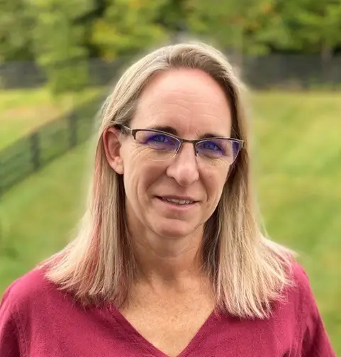 Smiling woman with shoulder-length blonde hair and glasses stands outdoors in a grassy area with trees.