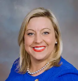 Smiling blonde woman in a blue top and pearl necklace, photographed against a soft gray background.