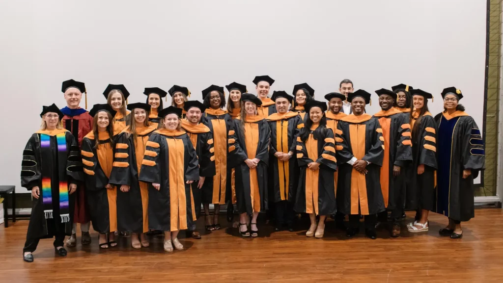 Graduates in caps and gowns stand smiling with faculty members, posed indoors against a plain background.