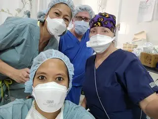 Four healthcare workers in scrubs, masks, and hair covers smile together in a medical setting.