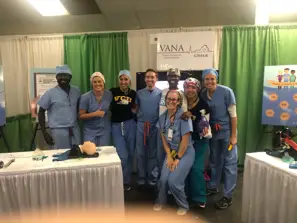 Eight smiling healthcare professionals stand together at a booth with medical equipment and banners in the background.