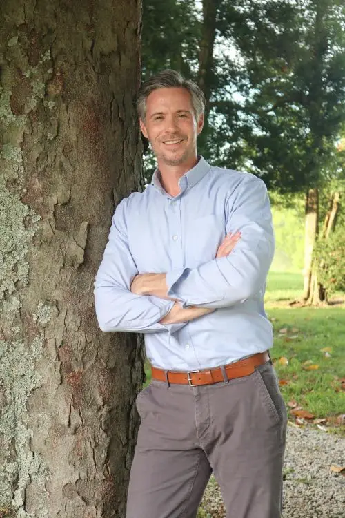 Smiling man with gray hair and light blue shirt stands outdoors, arms crossed, near a tree with greenery in the background.