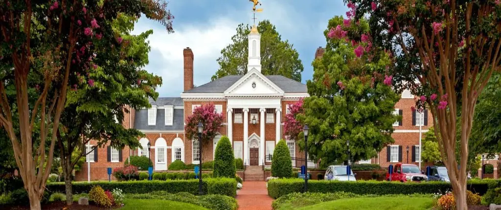 Colonial-style red-brick building with white columns, cupola, landscaped lawn, flowers, and trees under a partly cloudy sky.