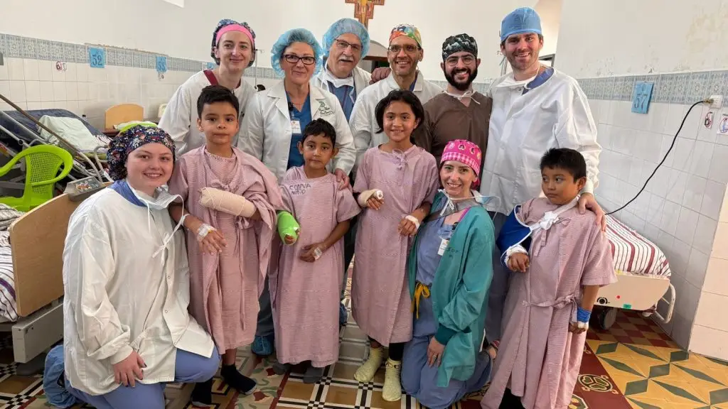 Doctors and nurses smile with four children in hospital gowns and arm casts, posing together in a hospital room.