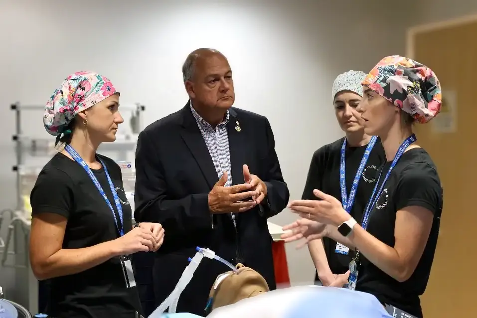 Three women in scrubs and a man in a suit discuss by a medical mannequin and equipment, suggesting collaborative training.