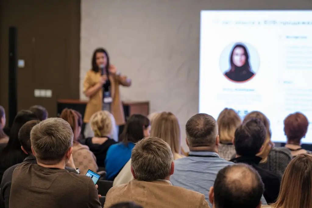 Woman addresses an audience at a conference, with a blurred presentation slide visible on the screen behind her.