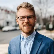 Smiling man with blond hair, glasses, and beard in a blue blazer outdoors with blurred houses and trees behind him.