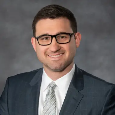 Smiling man in glasses, dark suit, and tie poses for a professional headshot with a gray background.