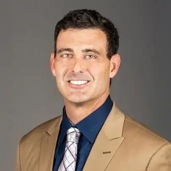 Smiling man in a tan suit jacket and navy shirt, posed against a gray background.