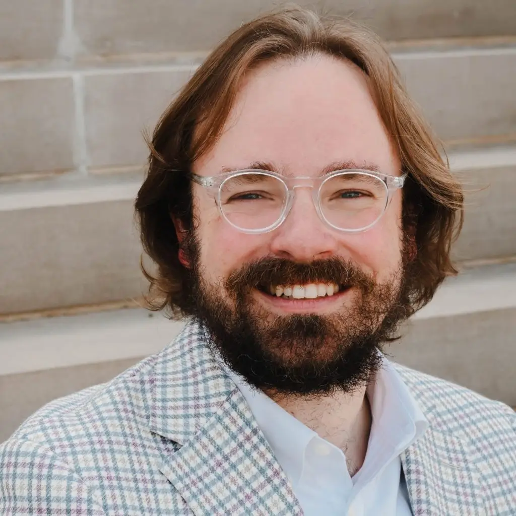 Smiling man with long brown hair, a beard, and glasses in a plaid jacket sits in front of stone steps.