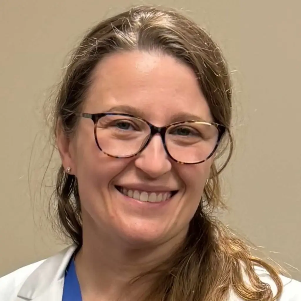 Smiling woman with long light brown hair, glasses, and a white coat stands before a beige background.